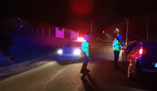 Două persoane purtând veste fluorescente stau pe un drum lângă mașini sub lumini colorate pe timp de noapte.