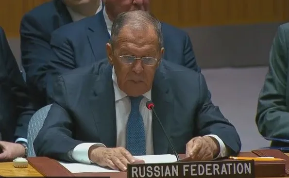 Un bărbat cu ochelari și costum închis se află așezat la o masă cu un semn pe care scrie RUSSIAN FEDERATION.