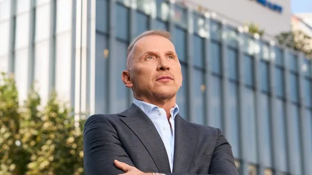 Un bărbat cu părul scurt și cămașă albastră sub un sacou gri stă cu brațele încrucișate în fața unei clădiri cu ferestre mari de sticlă.