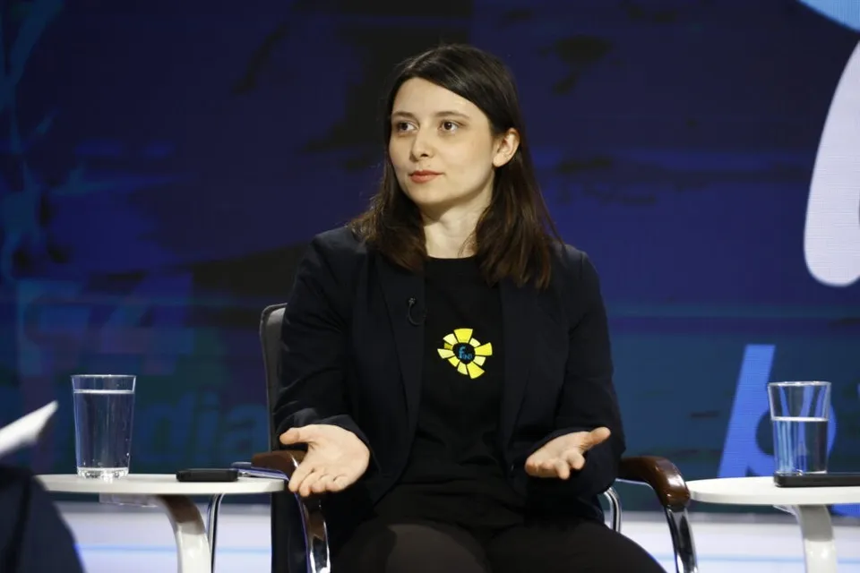 O femeie cu părul lung și întruchipată într-un sacou negru stă pe un scaun cu mâinile întinse în lateral.