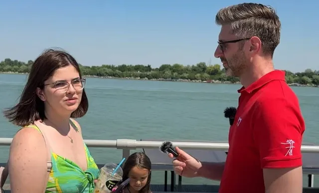 O femeie cu ochelari și o rochie verde stă lângă un bărbat cu tricou roșu care ține un microfon în fața unui fluviu.