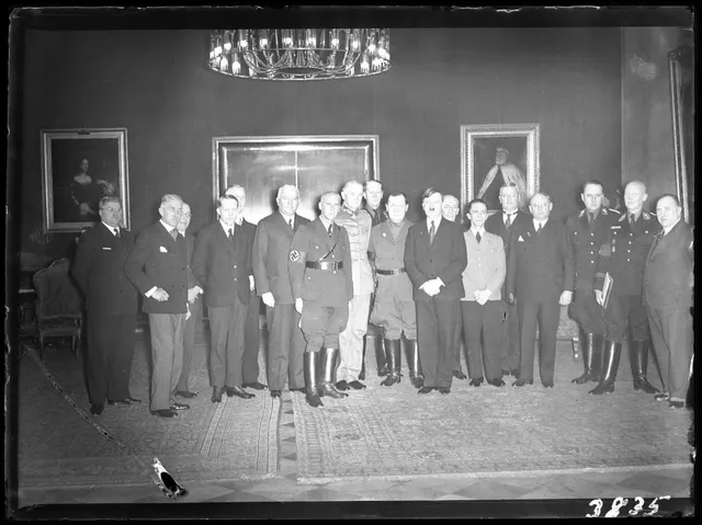 Grup de bărbați în costume și uniforme, aliniați într-o sală cu candelabru și tablouri pe pereți.