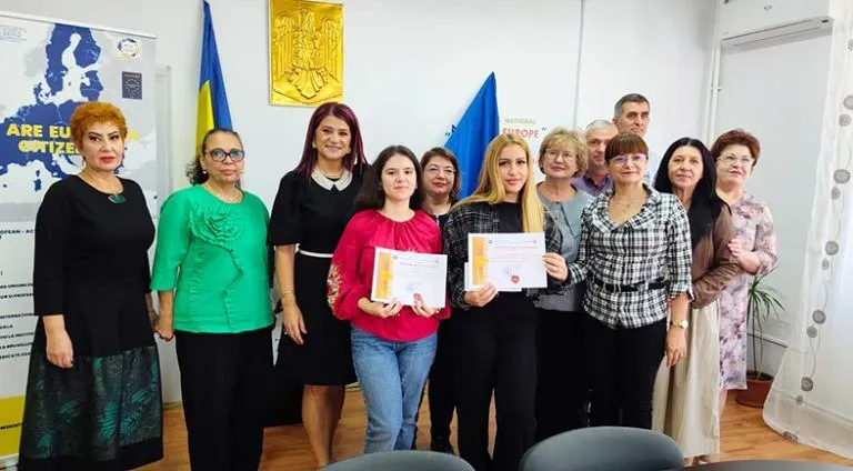 Un grup de femei stă în picioare într-o încăpere, unele purtând documente albe în mâini, în fața unui steag și al unui panou informativ.