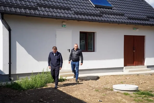 Doi bărbați merg pe un teren neamenajat în fața unei clădiri albe cu acoperiș închis și uși de lemn.