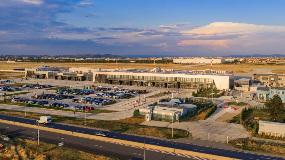O vedere aeriană a unei clădiri de terminal de aeroport cu o parcare plină de mașini și o autostradă în prim-plan.