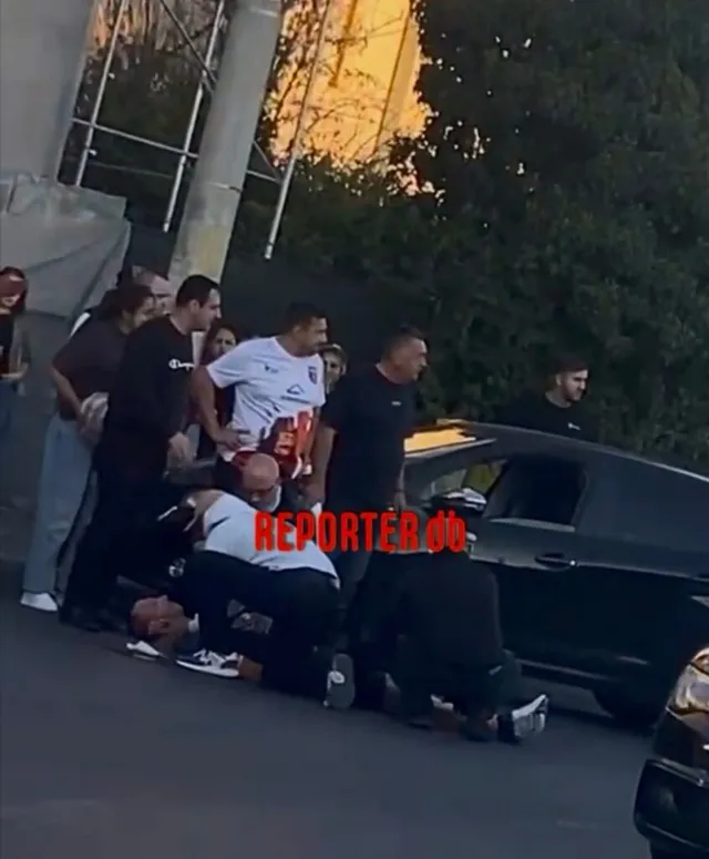 Mai mulți bărbați stau în jurul unei persoane întinse pe asfalt lângă o mașină neagră.