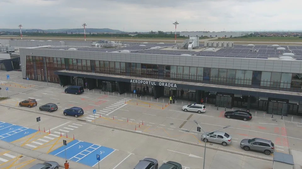 O clădire modernă cu ferestre mari, un parc auto în prim-plan și panouri solare instalate pe acoperișul unei structuri lungi.