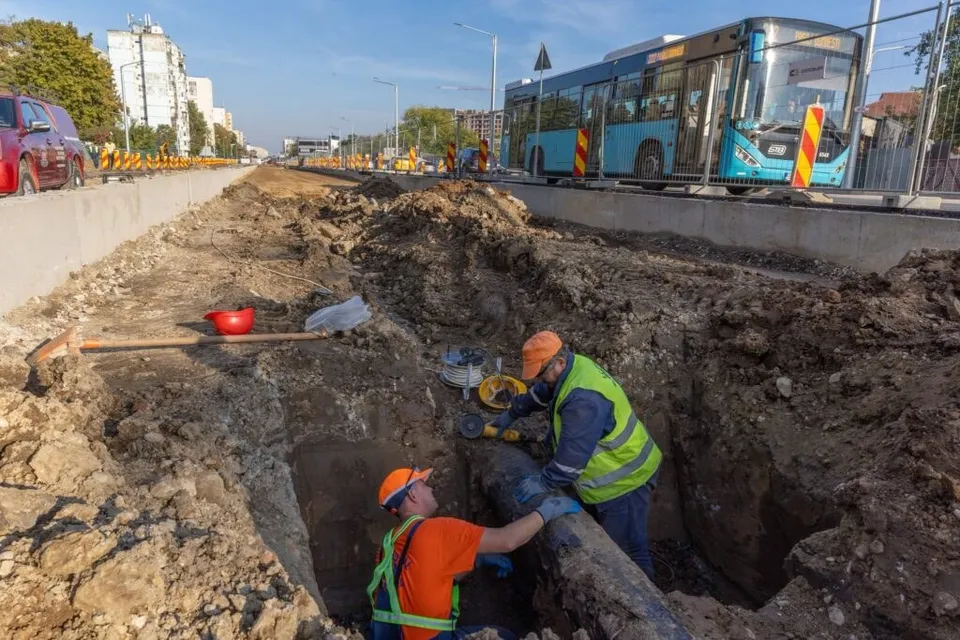 Unii muncitori cu căști portocalii și veste reflectorizante lucrează într-o șanț adânc de pământ lângă o stradă cu un autobuz albastru.