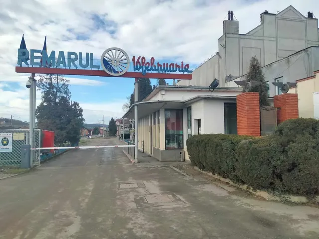 O poartă metalică susținută de stâlpi gri poartă un panou cu textul REMARUL 16 februarie deasupra unui drum de asfalt.