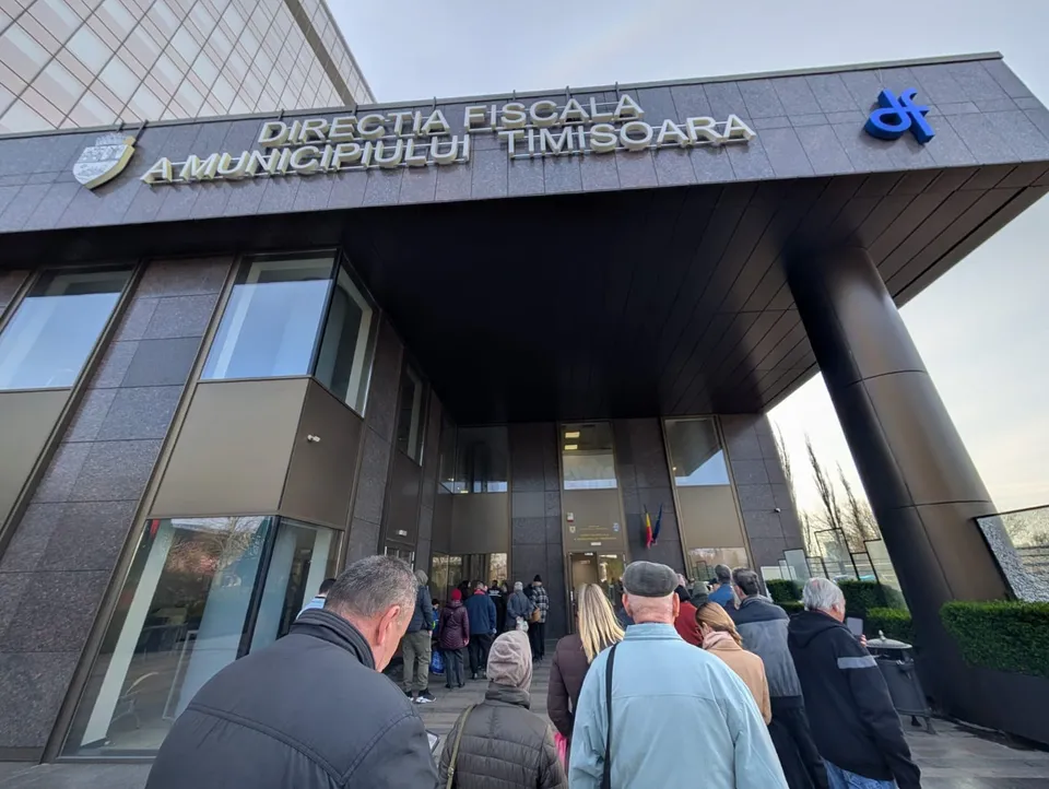 O mulțime de oameni stă la coadă în fața unei clădiri moderne cu panouri de sticlă și un panou de identificare pe fațadă.