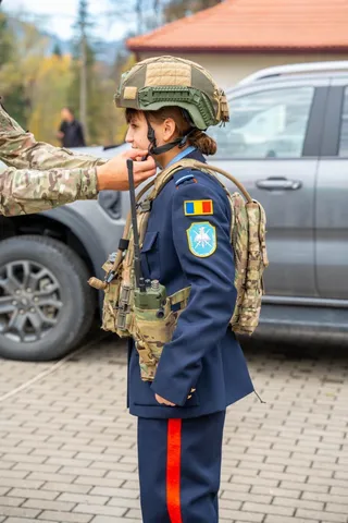 O persoană în uniformă albastră cu un rucsac camuflaj și cască militară stă lângă un vehicul gri.