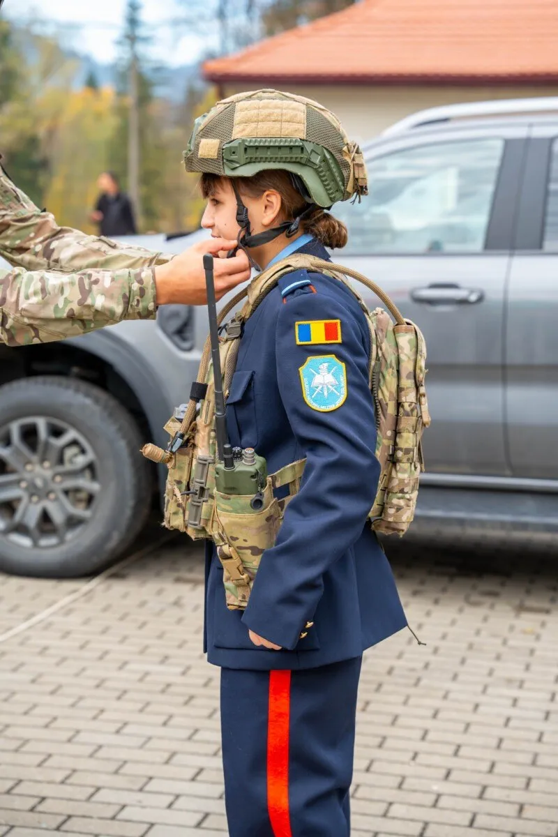 O persoană în uniformă albastră cu un rucsac camuflaj și cască militară stă lângă un vehicul gri.