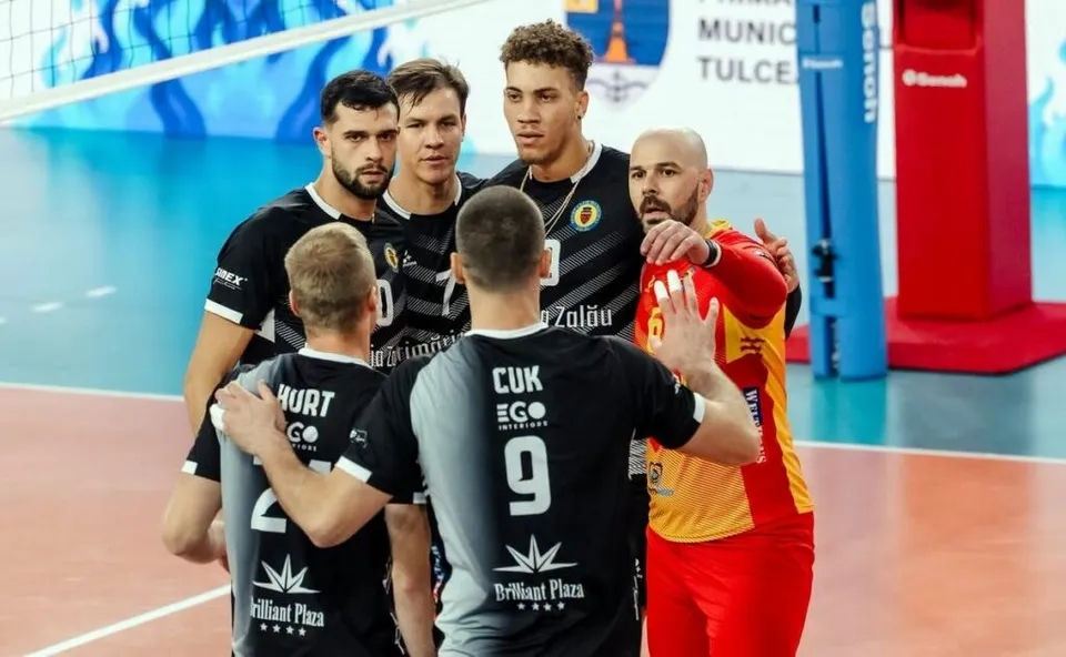 Cinci jucători de volei în echipamente negre și unul în echipament roșu și galben stau grupați pe un teren de sport.
