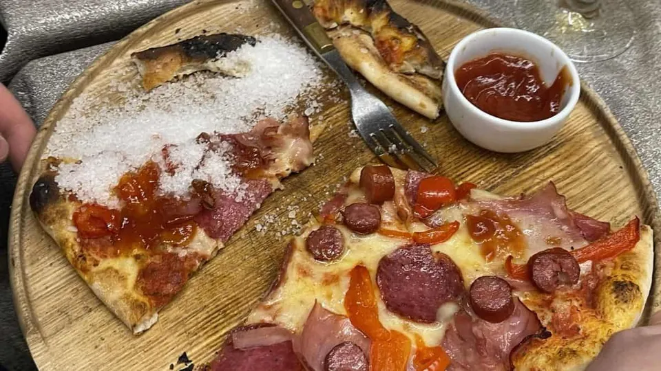 Două felii de pizza cu diverse ingrediente și un bol mic cu sos roșu sunt așezate pe o farfurie de lemn rotundă.