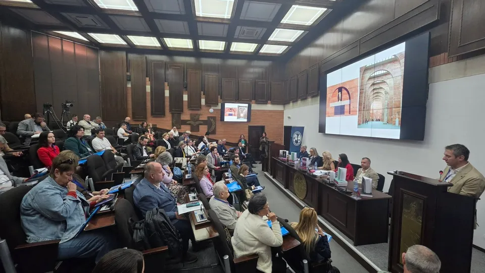 Oamenii stau așezați în scaunele unui auditorium, privind spre un podium și un ecran mare care afișează o imagine cu o poartă de piatră.