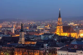 Panorama unui oraș iluminat la seară cu clădiri diverse și un turn de biserică înalt sub un cer de seară.