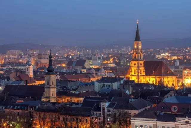 Panorama unui oraș iluminat la seară cu clădiri diverse și un turn de biserică înalt sub un cer de seară.