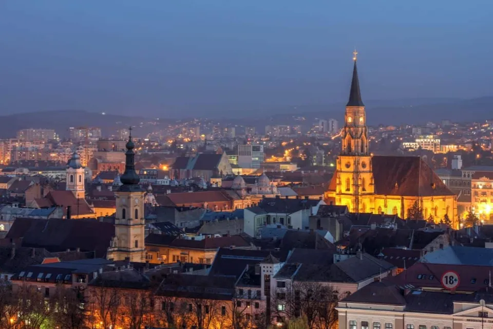Panorama unui oraș iluminat la seară cu clădiri diverse și un turn de biserică înalt sub un cer de seară.