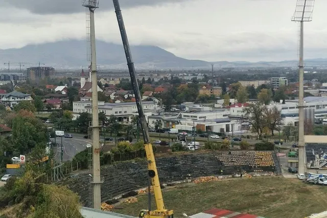 O macararie galbenă este poziționată lângă tribunele unui stadion, sub un cer înnorat, având clădiri și munți în fundal.