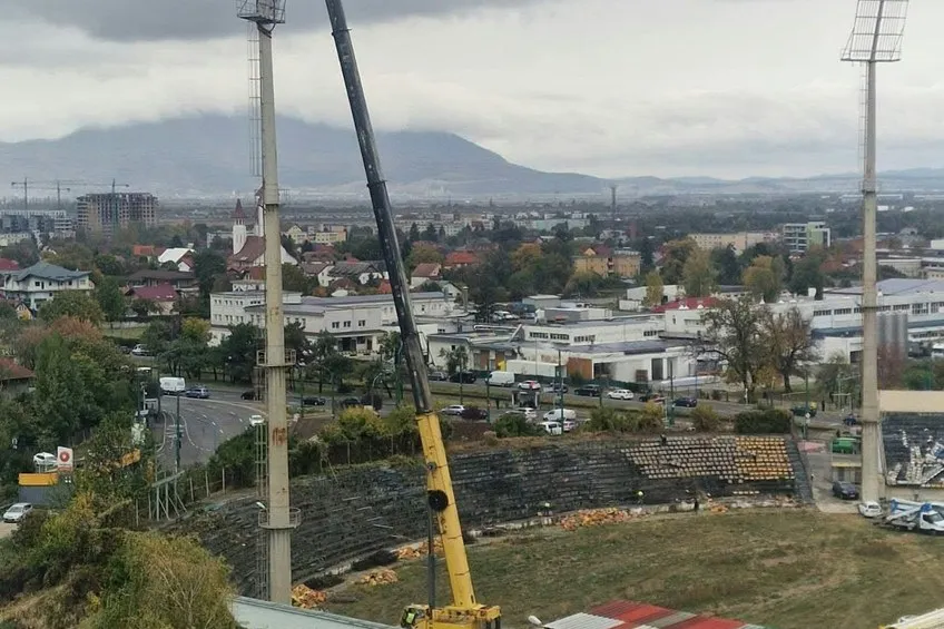 O macararie galbenă este poziționată lângă tribunele unui stadion, sub un cer înnorat, având clădiri și munți în fundal.