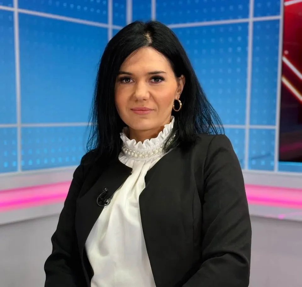 O femeie cu părul negru, purtând un sacou negru peste o bluză albă cu guler dantelat, privește spre cameră într-un studio cu fundal albastru.