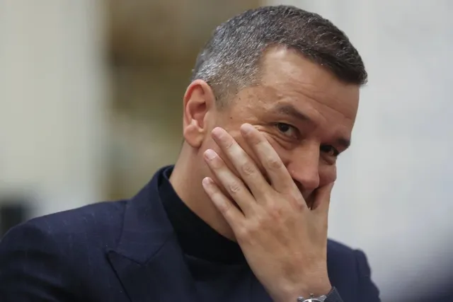 Un bărbat cu părul scurt și scurtat pe lateral poartă un sacou în dungi și un tricou cu guler înalt, având mâna pe față.