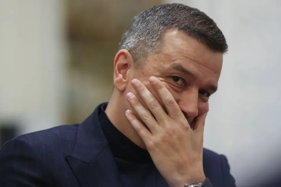 Un bărbat cu părul scurt și scurtat pe lateral poartă un sacou în dungi și un tricou cu guler înalt, având mâna pe față.