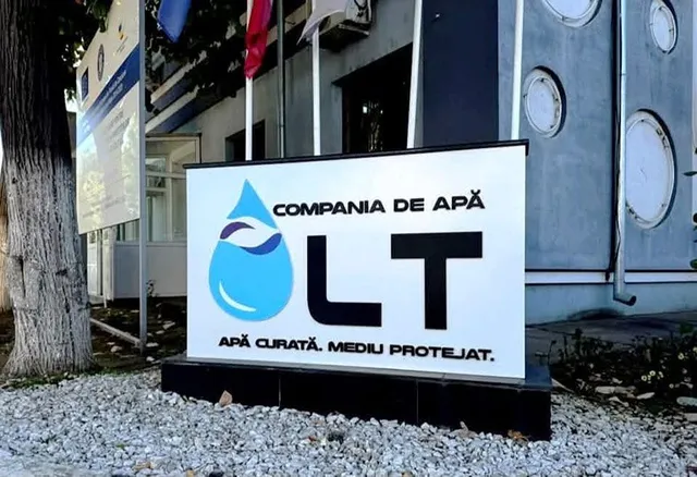 Un semn alb cu textul Compania de Apă Olt și un logo albastru în formă de picătură este amplasat pe un suport negru lângă o clădire.