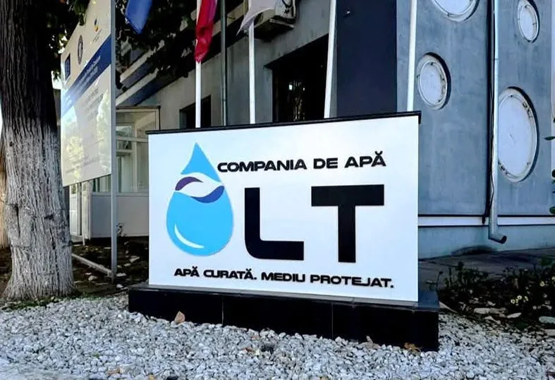Un semn alb cu textul Compania de Apă Olt și un logo albastru în formă de picătură este amplasat pe un suport negru lângă o clădire.