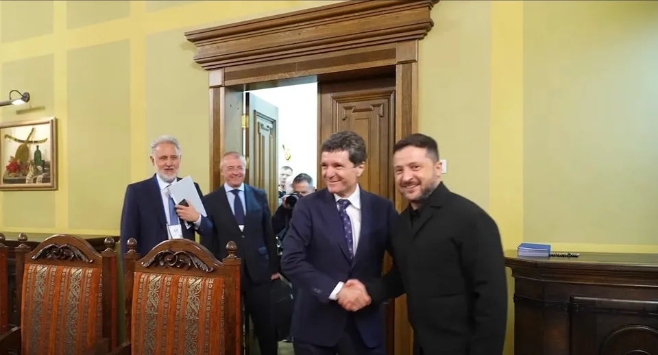 Patru bărbați în costume și haine închise stau în picioare într-o încăpere cu pereți galbeni și uși din lemn, unul dintre ei zâmbind spre cameră.