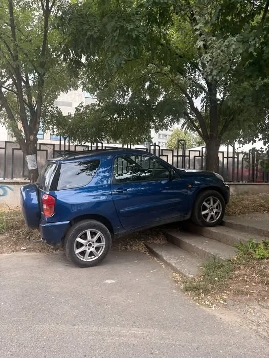 Un autoturism albastru este parcat pe treptele de beton dintre o zonă asfaltată și un gard metalic, sub coroana unor copaci verzi.