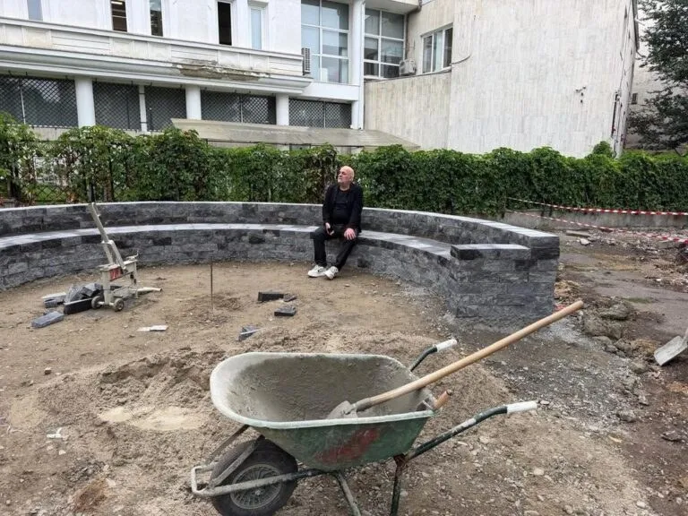 Un bărbat stă așezat pe un zid curb din piatră într-un șantier cu un cărucior de sârmă și un utilaj de tăiat în prim-plan.