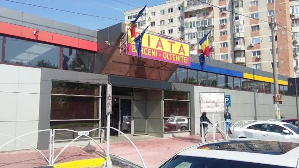 O clădire modernă cu ferestre mari, un panou publicitar colorat și câteva mașini parcate în fața acesteia sub un cer senin.