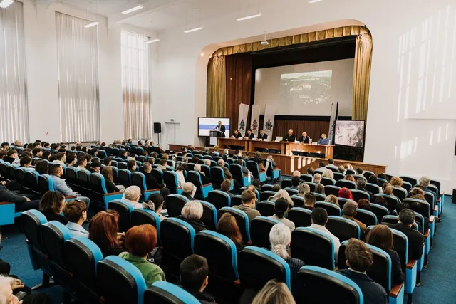 O mulțime de oameni stă așezată în scaune de culoare albastră într-o sală de conferințe cu un podium și un ecran de proiecție în față.