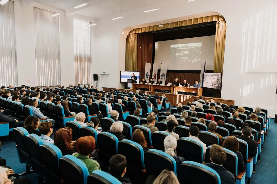 O mulțime de oameni stă așezată în scaune de culoare albastră într-o sală de conferințe cu un podium și un ecran de proiecție în față.