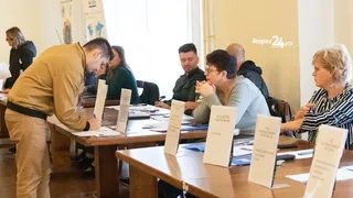 Un bărbat în cămașă galbenă se apleacă peste o masă de lemn, în timp ce mai mulți oameni stau așezați la mese cu diverse documente și pliante albe.