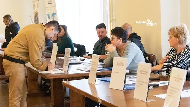 Un bărbat în cămașă galbenă se apleacă peste o masă de lemn, în timp ce mai mulți oameni stau așezați la mese cu diverse documente și pliante albe.