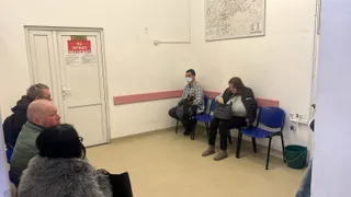 Câteva persoane stau pe scaune într-o încăpere cu pereți albi, având o ușă și un poster pe perete.