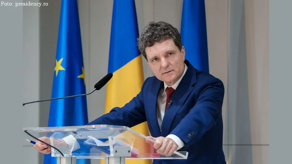 Un bărbat cu sacou albastru și cravată roșie stă în spatele unui podium transparent în fața unor steaguri albastre și galbene.