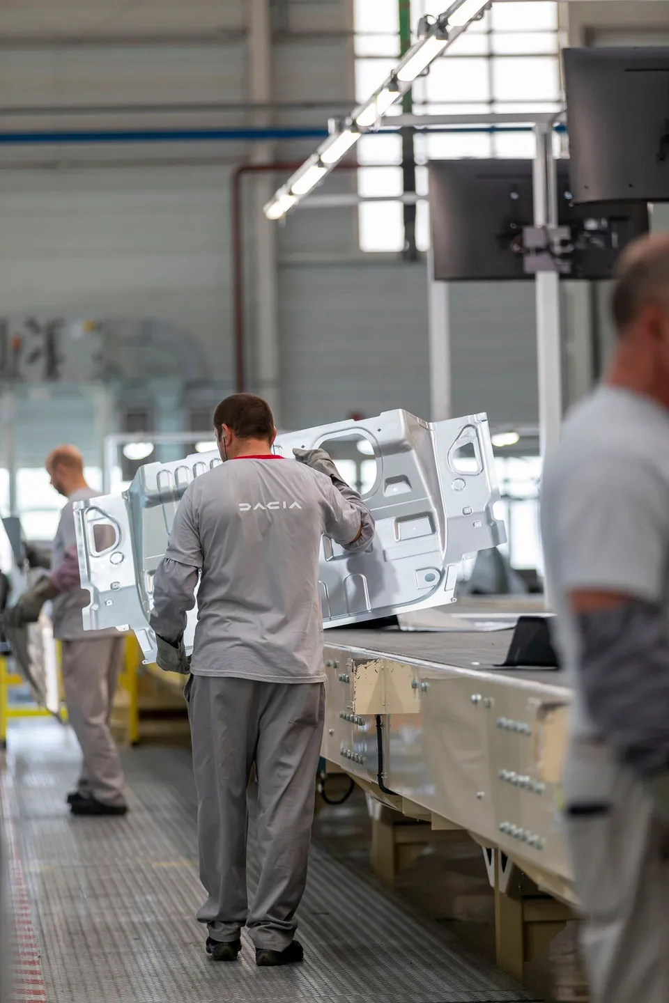 Un bărbat cu tricou gri și pantaloni de lucru gri manipulează o piesă metalică albă pe o linie de producție într-o fabrică.