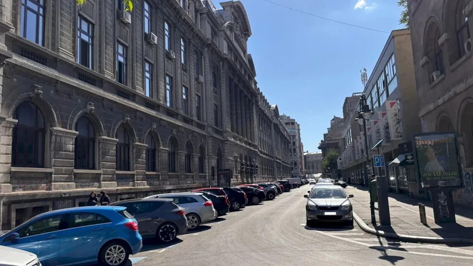 O stradă urbană cu mașini parcate pe partea stângă și un autoturism gri circulând pe asfaltul unei căi pietonale sau străzi.