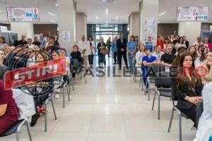 Un grup de persoane stă așezat pe scaune dispuse în două rânduri, într-un hol cu podea deschisă și pereți albi.