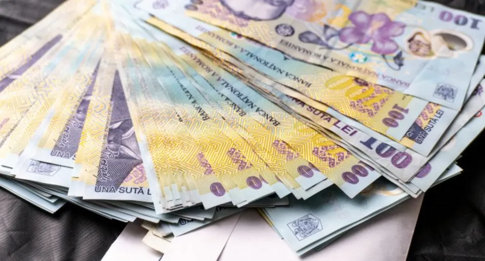 Un teanc de bancnote românești de o sută de lei este întins pe o suprafață întunecată.