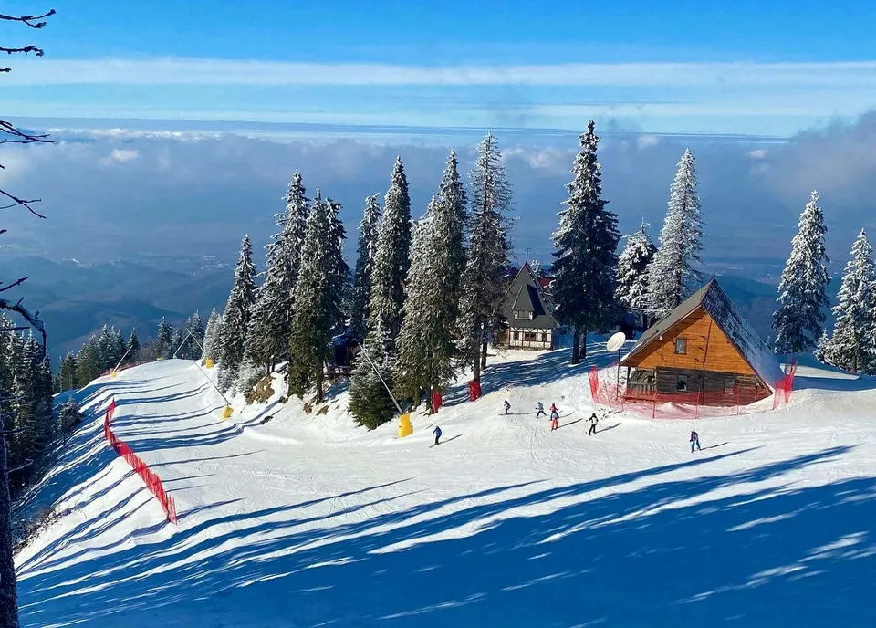 Oamenii schiază pe o pârtie de schi împădurită cu conifere acoperite de zăpadă, lângă câteva cabane de lemn sub un cer senin.