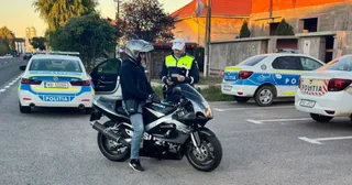 Un agent de poliție în uniformă cu vestă reflectorizantă stă lângă un motociclist purtând cască pe o motocicletă neagră, în fața unor mașini de poliție.