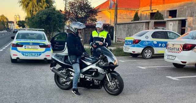 Un agent de poliție în uniformă cu vestă reflectorizantă stă lângă un motociclist purtând cască pe o motocicletă neagră, în fața unor mașini de poliție.