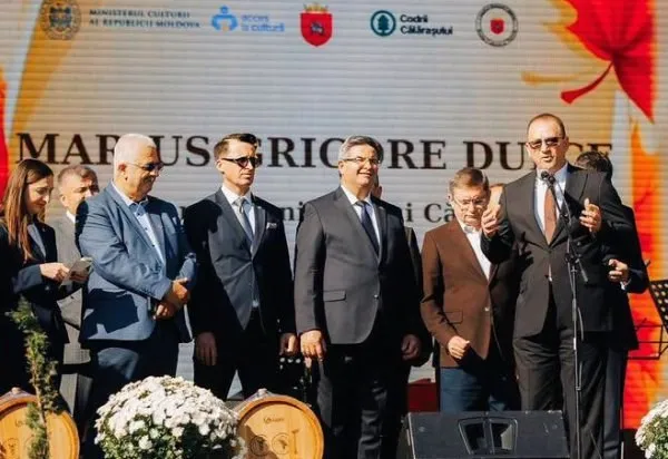 Un grup de bărbați în costume de sacou stă pe o scenă în fața unui panou publicitar cu diverse embleme și text.