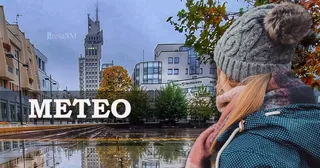 O persoană cu o căciulă gri și o jachetă albastră stă cu spatele, privind spre clădiri și copaci sub un cer înnorat.