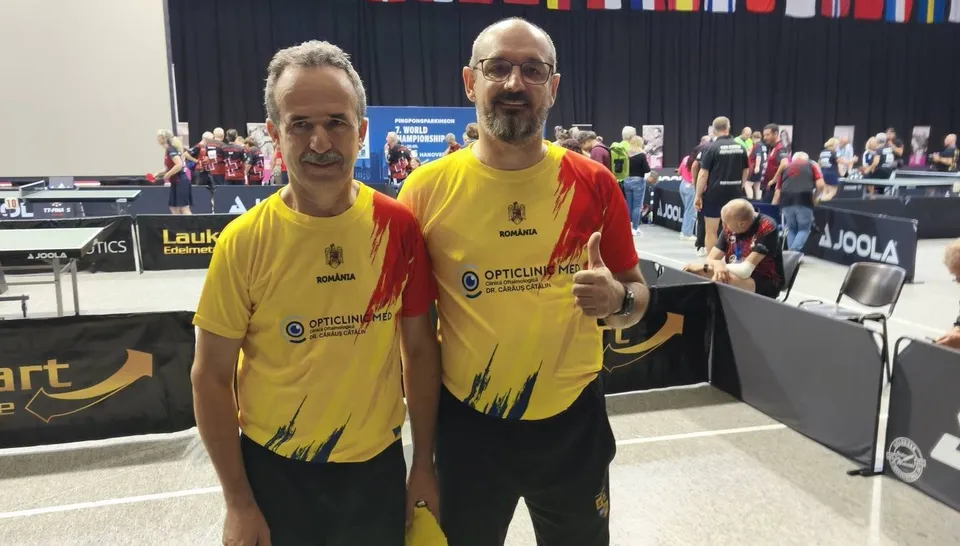 Doi bărbați îmbrăcați în tricouri galbene cu roșu stau în picioare într-o sală de sport cu mese de tenis de masă în fundal.