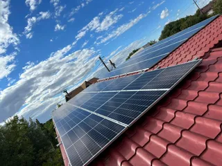 Panouri solare sunt instalate pe un acoperiș de țiglă roșie sub un cer albastru cu nori albi.